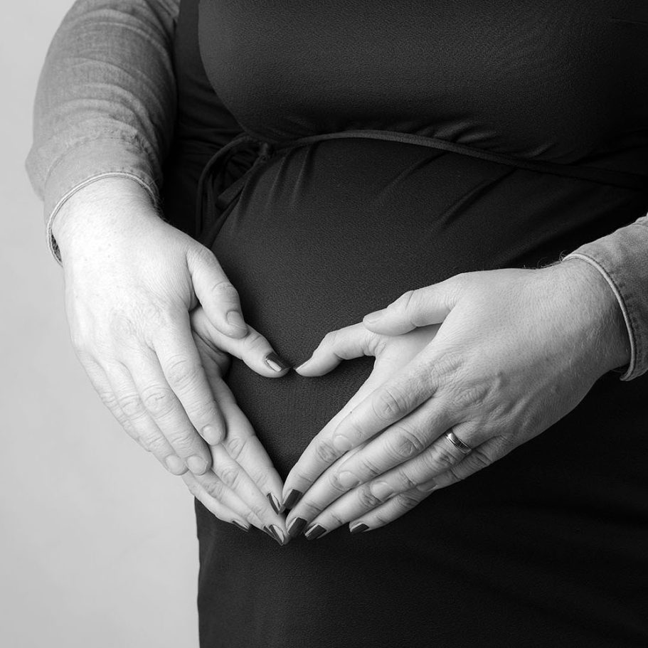 Detailaufnahme eines Babybauchs: Die Hände des Partners bilden ein Herz. Emotionales Schwangerschaftsshooting im Fotostudio Würzburg.